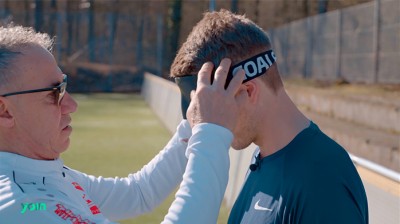 Trainer (links) setzt Spieler (rechts) Maske zur Verblindung auf. Aufnahme von der Seite zeigt beide vom Kopf bis zur Brust.